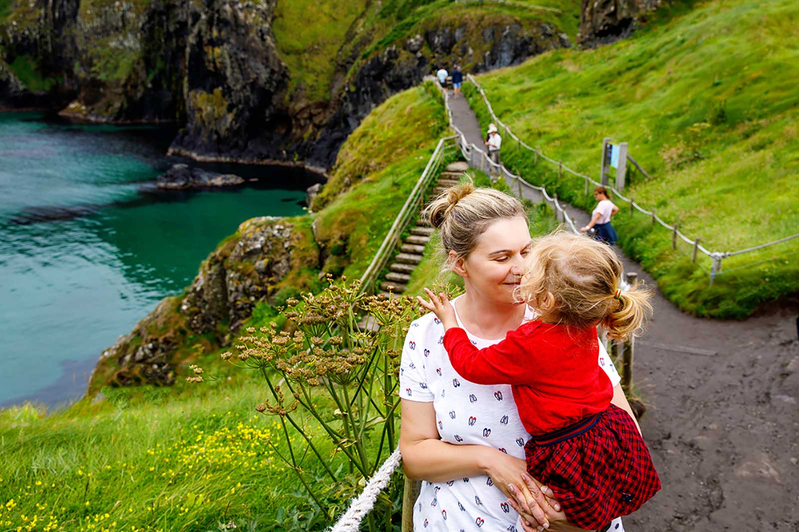 A scenic view of a coastal path with a woman holding a child. Lush green hills surround the turquoise water.