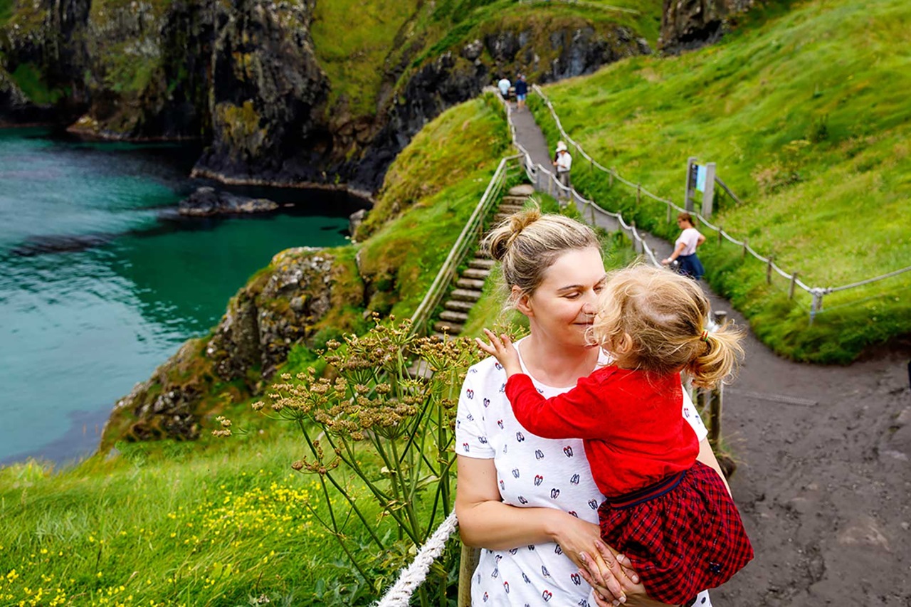 A scenic view of a coastal path with a woman holding a child. Lush green hills surround the turquoise water.