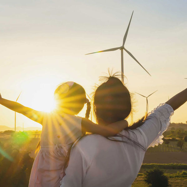 A mother and child embrace during sunset, with wind turbines in the background, symbolising nature and family connection.