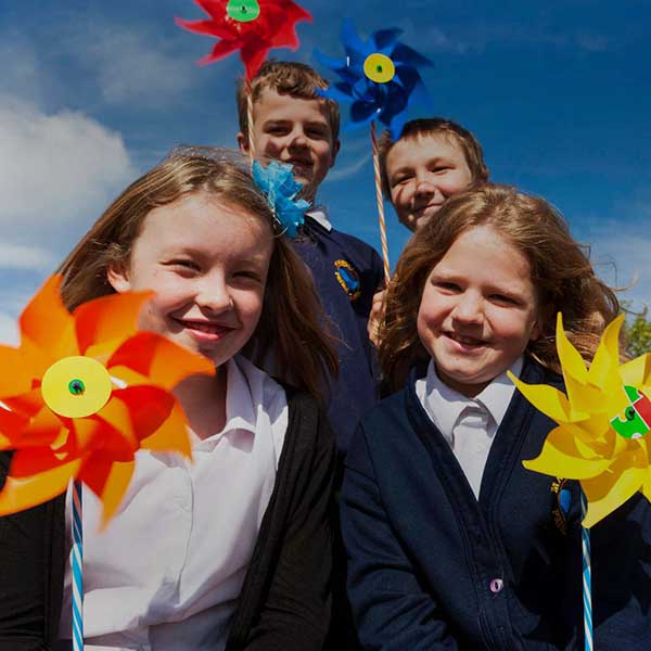 A group of children hold colourful pinwheels against a bright blue sky, smiling and dressed in school uniforms.