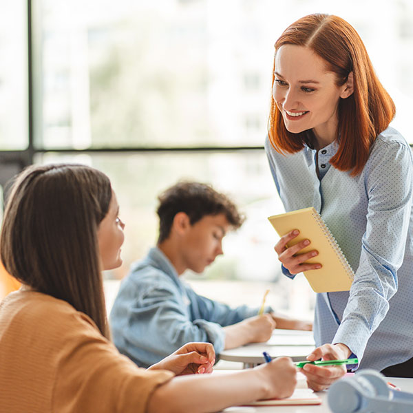 A teacher is assisting students at a desk, holding a notebook while they write and study together in a bright classroom.