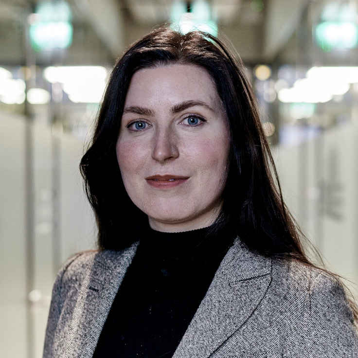 A woman in a grey blazer and black top stands in a modern indoor setting with blurred background elements.