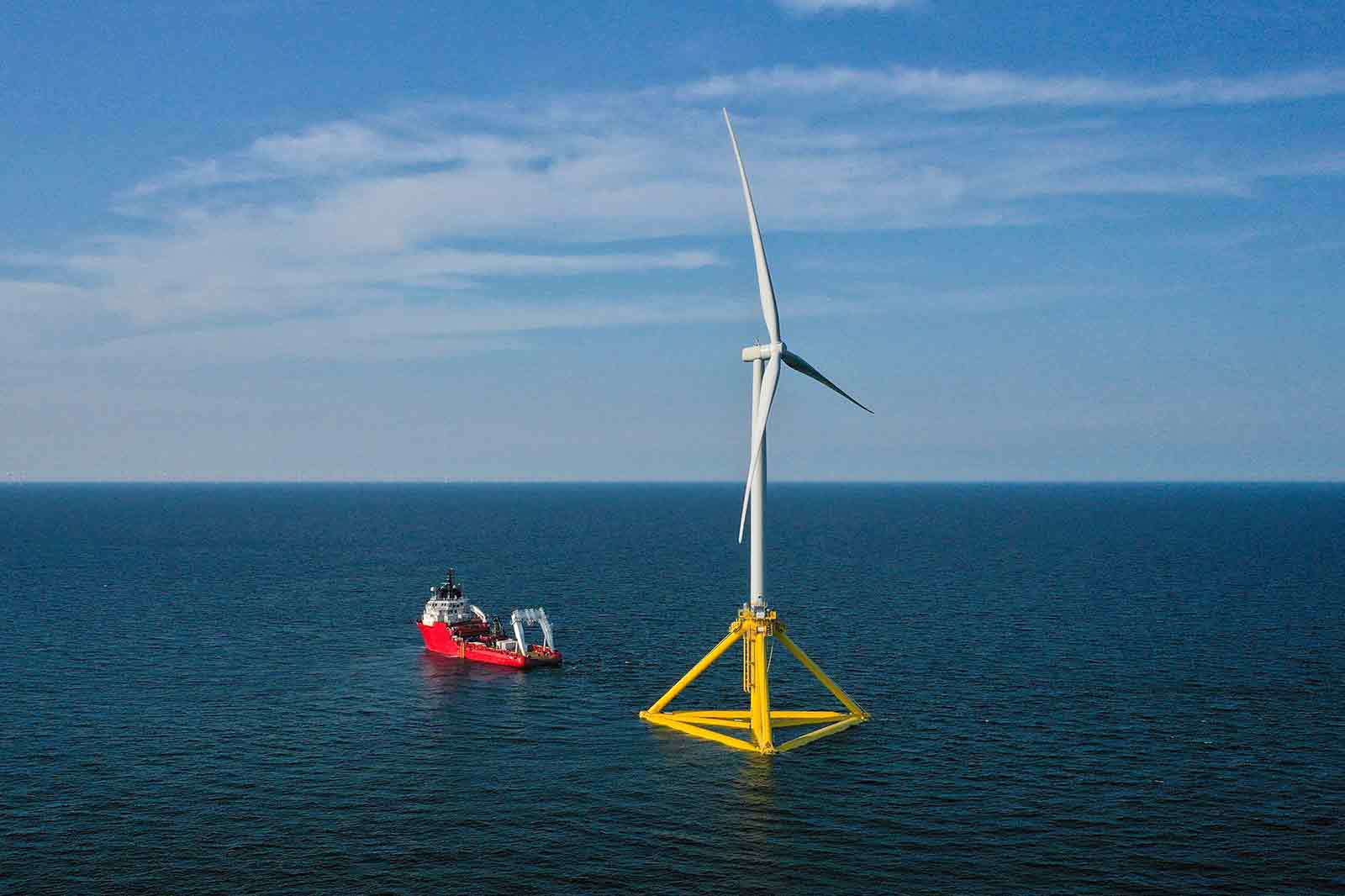 An offshore wind turbine with a yellow base stands in the sea. A red support vessel is nearby under a clear blue sky.