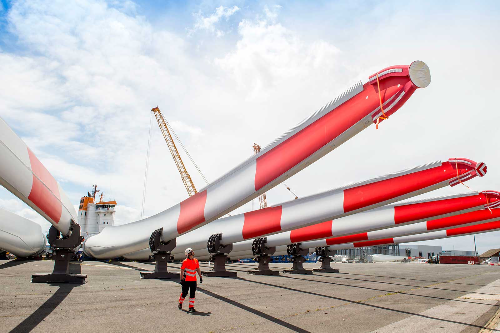 A worker in a red and white outfit stands near large, striped wind turbine blades at a dock with a crane and ship in the background.