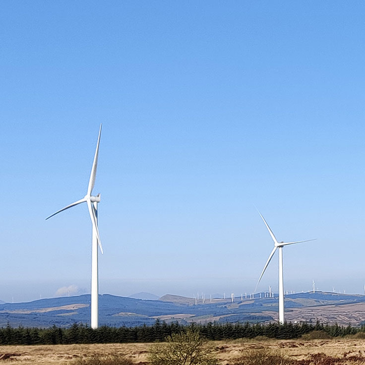 Two wind turbines stand against a clear blue sky, with mountains in the background and trees in the foreground.