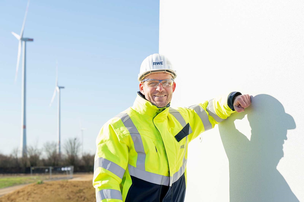 A person wearing a bright yellow safety jacket and a hard hat stands against a white wall, with wind turbines in the background.