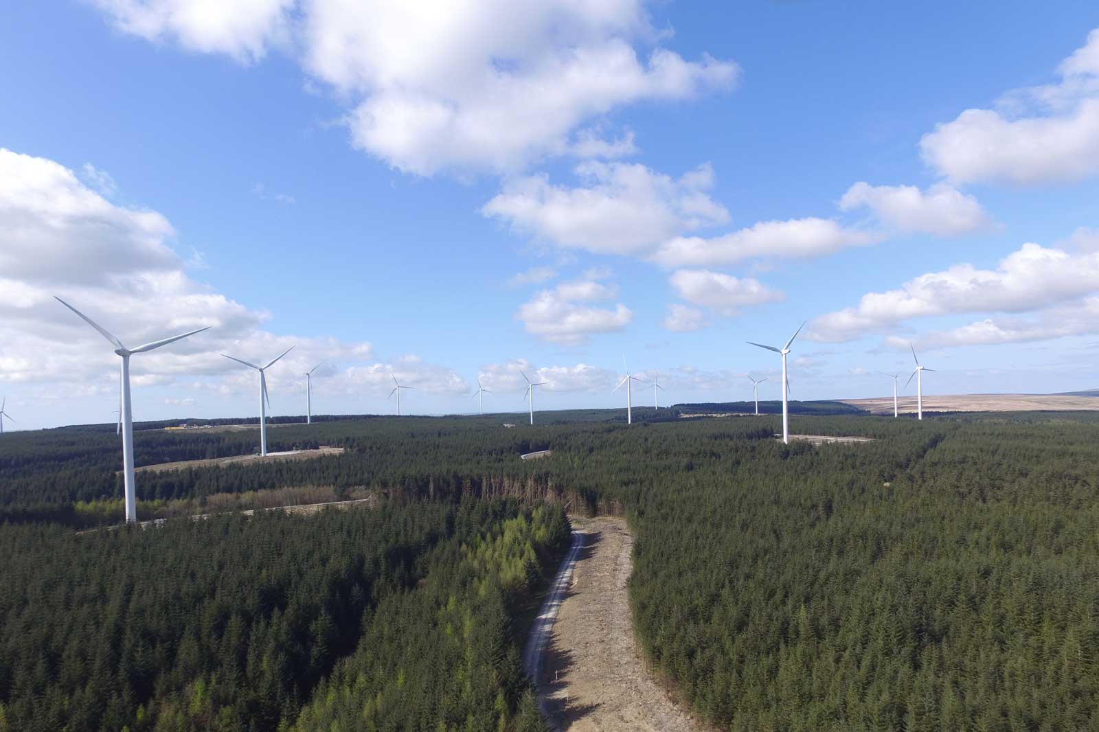Aerial view of a green forest landscape with several wind turbines under a blue sky with white clouds.