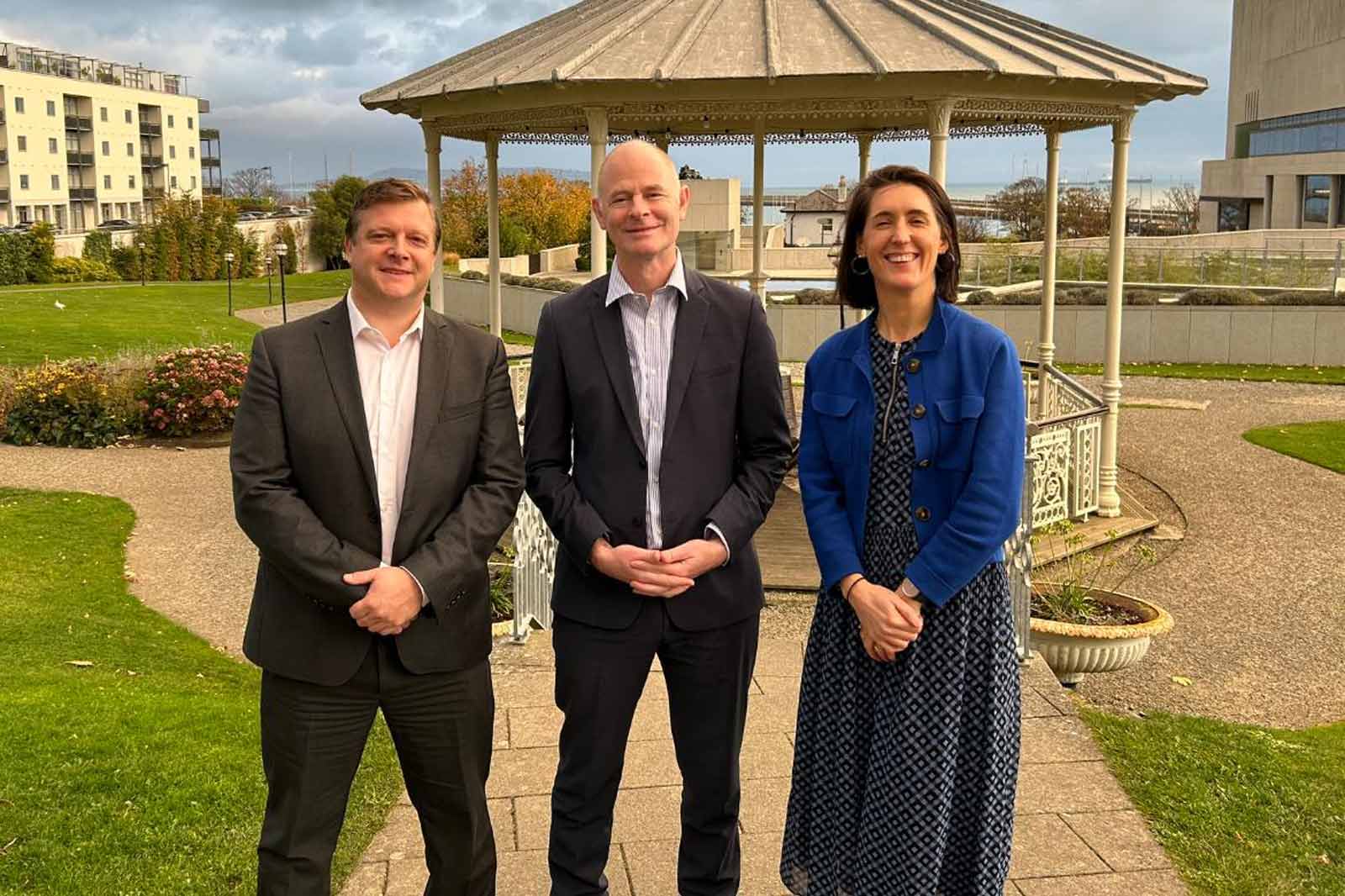 Three people stand in a park in front of a gazebo. Two men wear suits, and a woman is dressed in a blue coat.