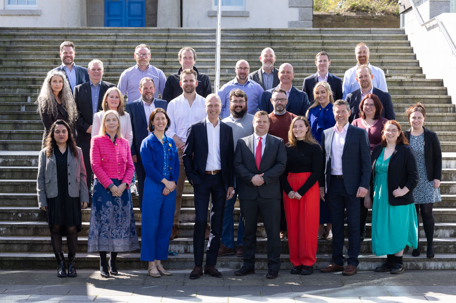 A group of diverse individuals posing on stairs, wearing formal and semi-formal attire in an outdoor setting.