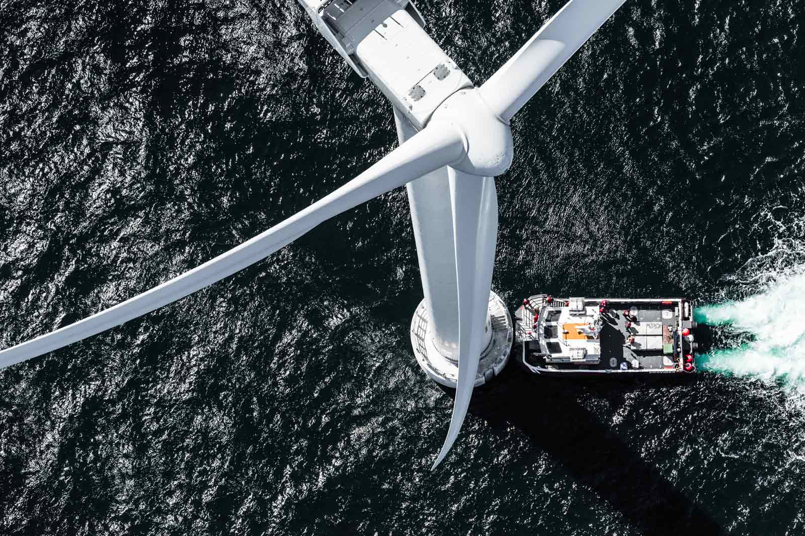 A bird's-eye view of a large offshore wind turbine, with a service boat beneath it on the dark, turbulent water.