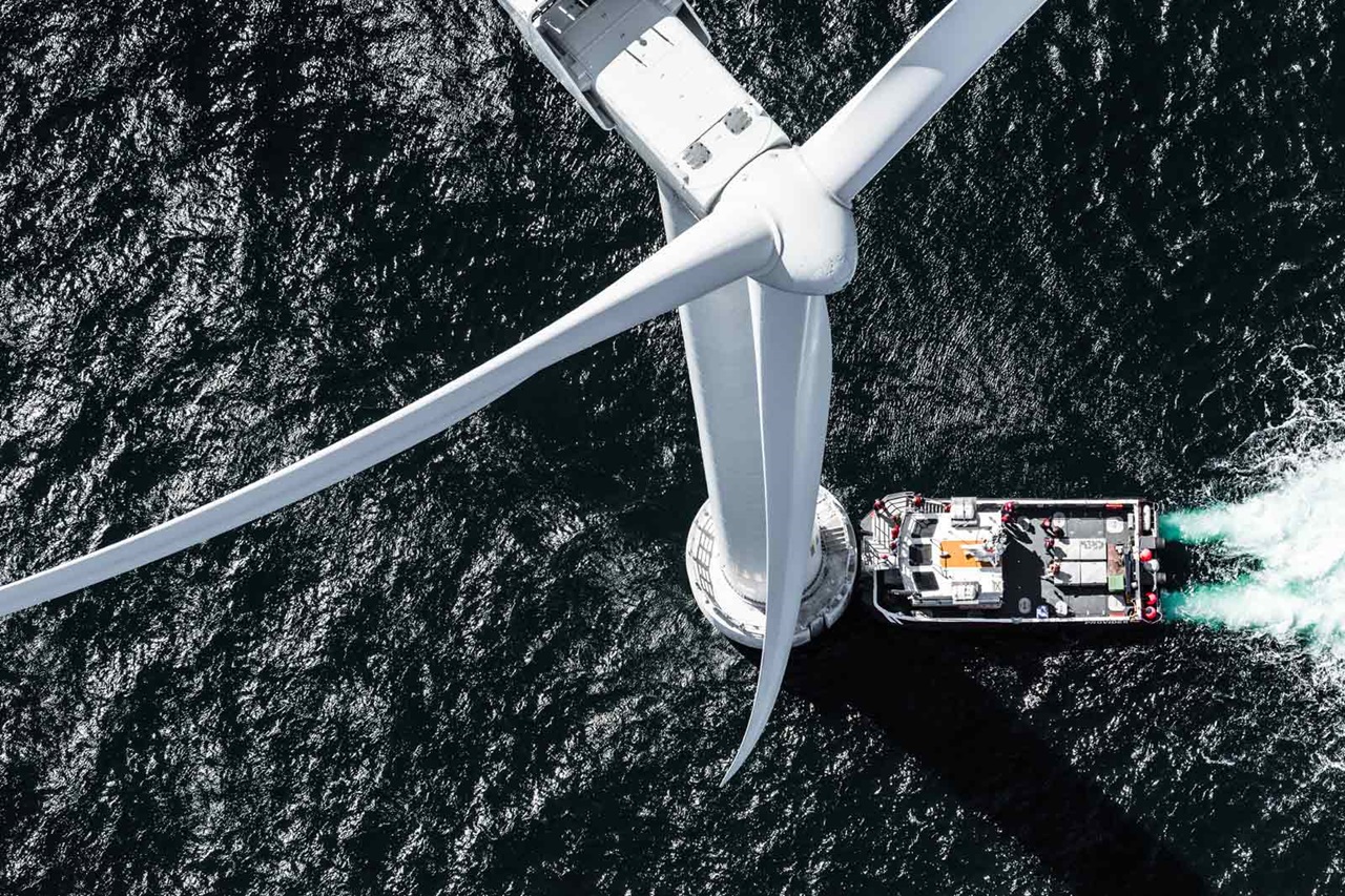 A bird's-eye view of a large offshore wind turbine, with a service boat beneath it on the dark, turbulent water.