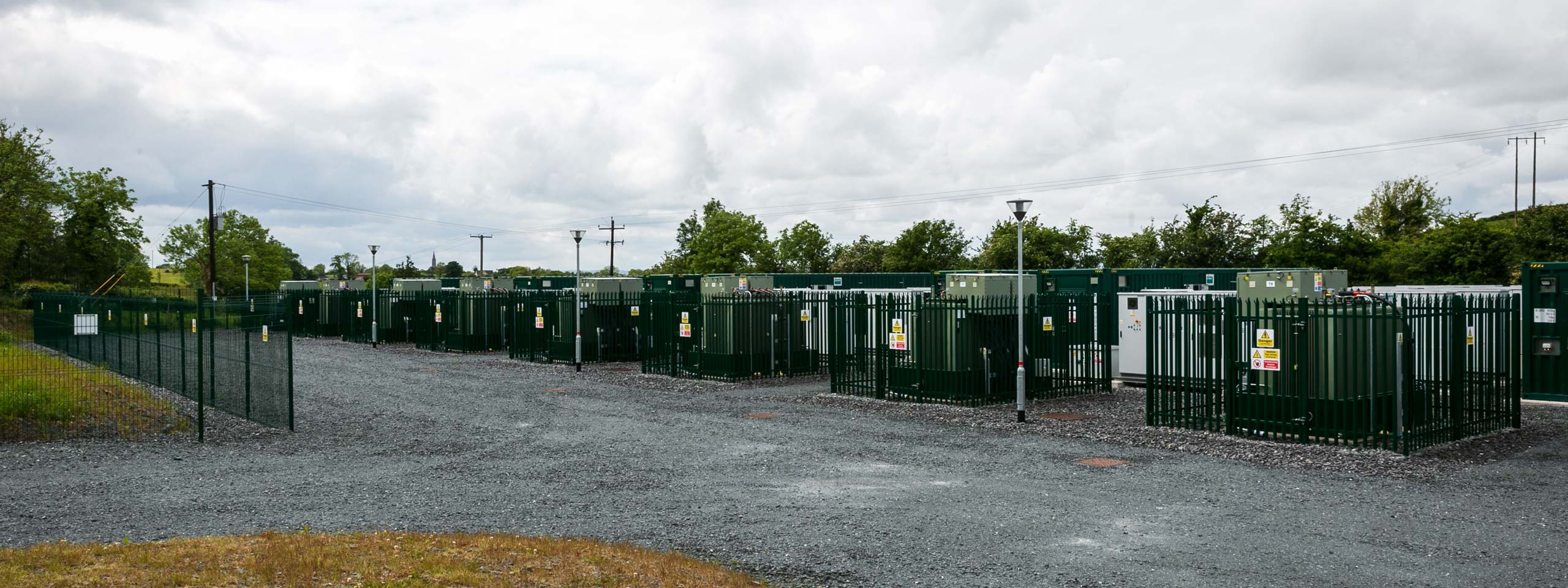 A view of multiple green storage containers fenced in a gravel area, with trees and a cloudy sky in the background.