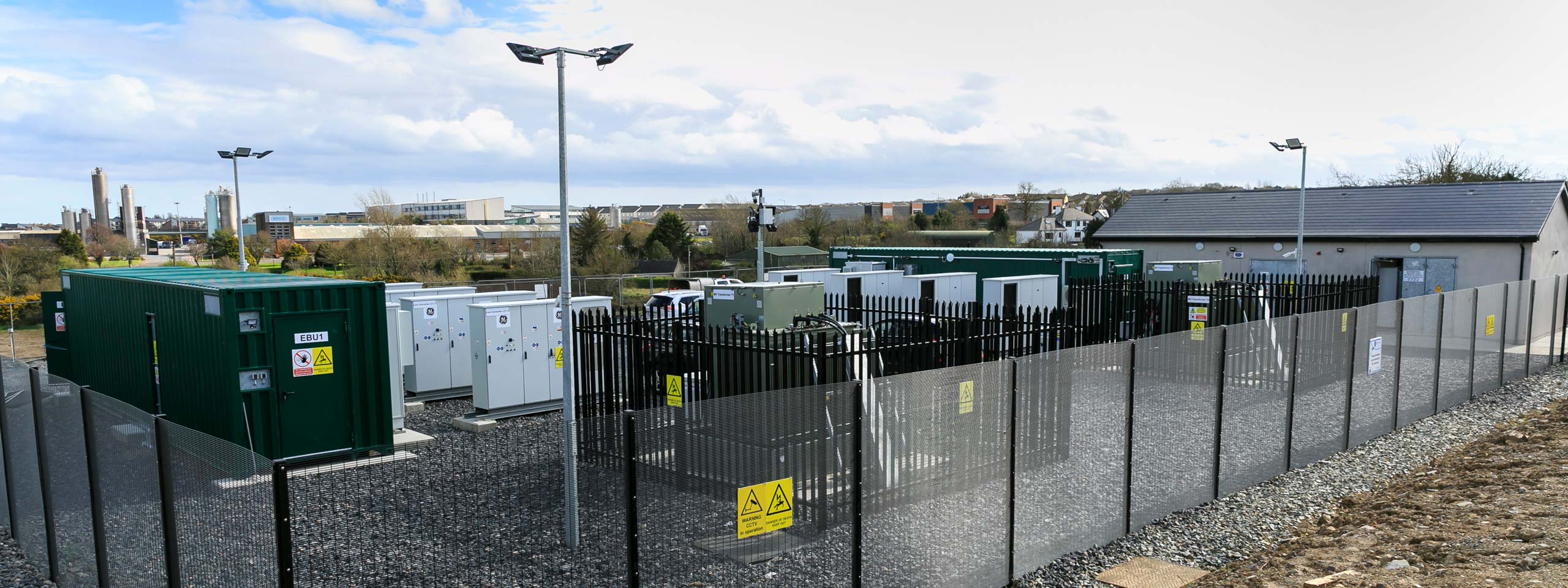 A secured facility with green shipping containers, electrical units, and fencing, located under a cloudy sky.