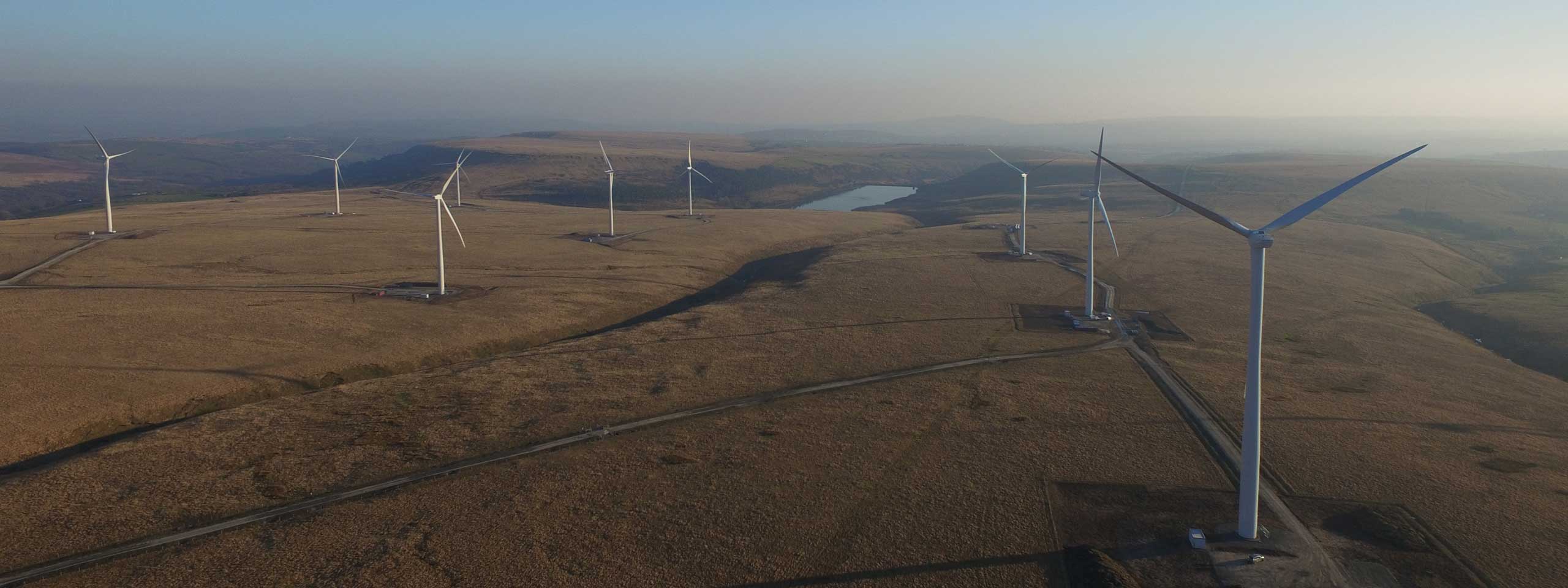 Aerial view of multiple wind turbines on a grassy hillside with a lake in the background under a clear sky.