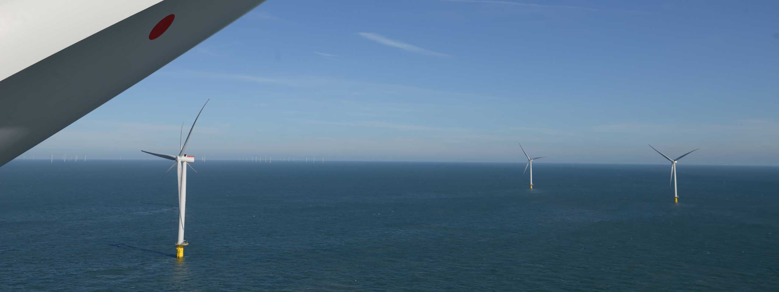 A view of offshore wind turbines in the sea under a clear blue sky, with a wind turbine blade partially visible.