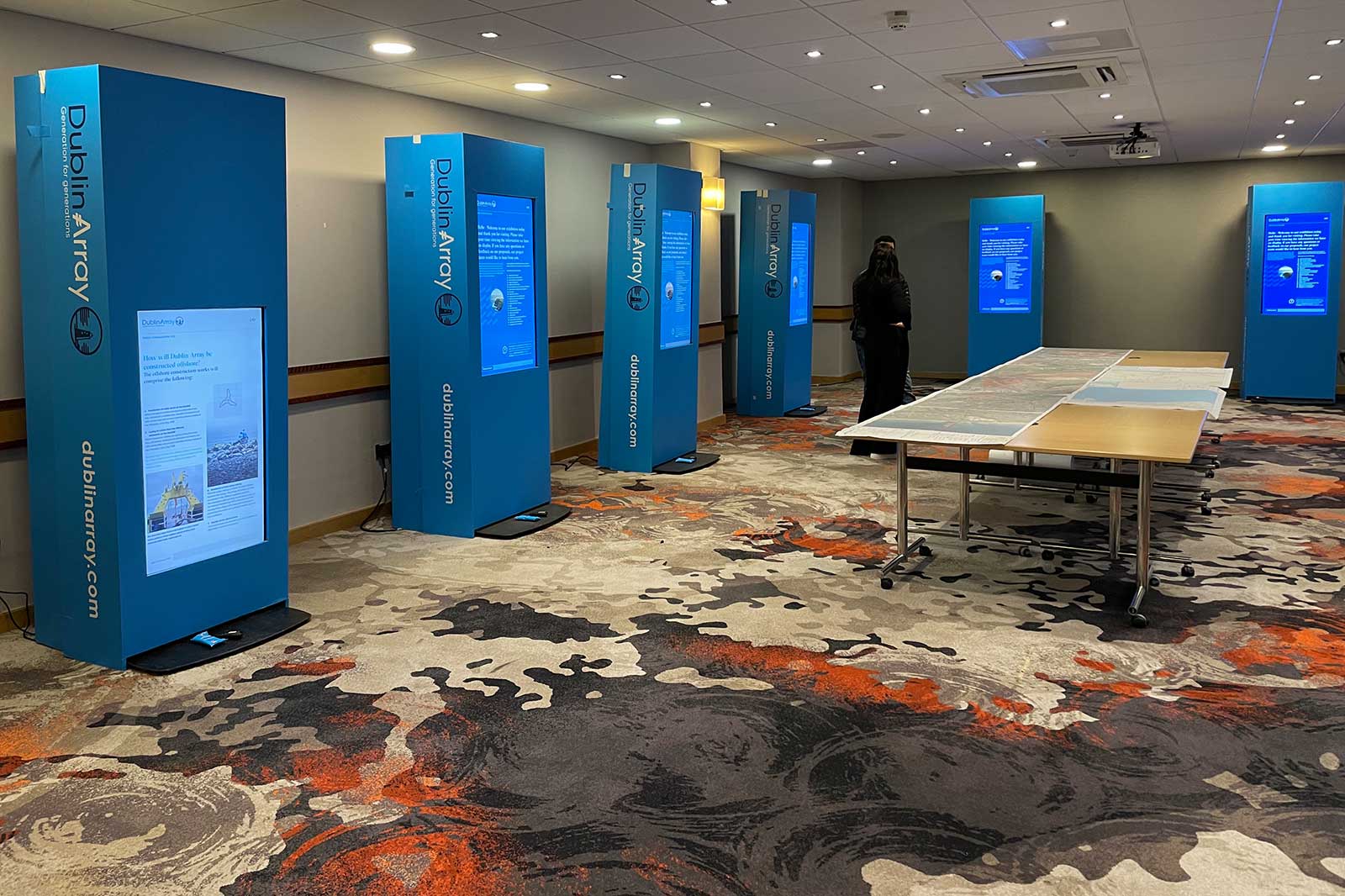 A conference room featuring interactive displays and tables with maps. A person stands near the displays.