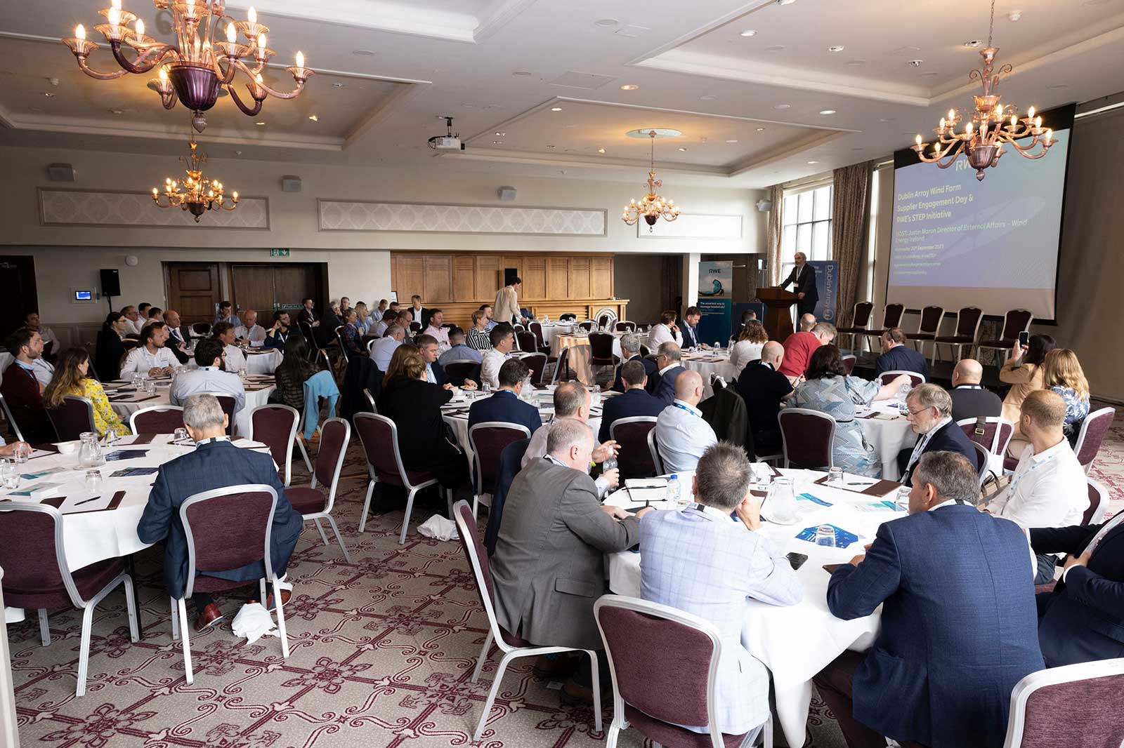 A conference room filled with attendees seated at tables, listening to a speaker presenting on a screen in a well-lit setting.