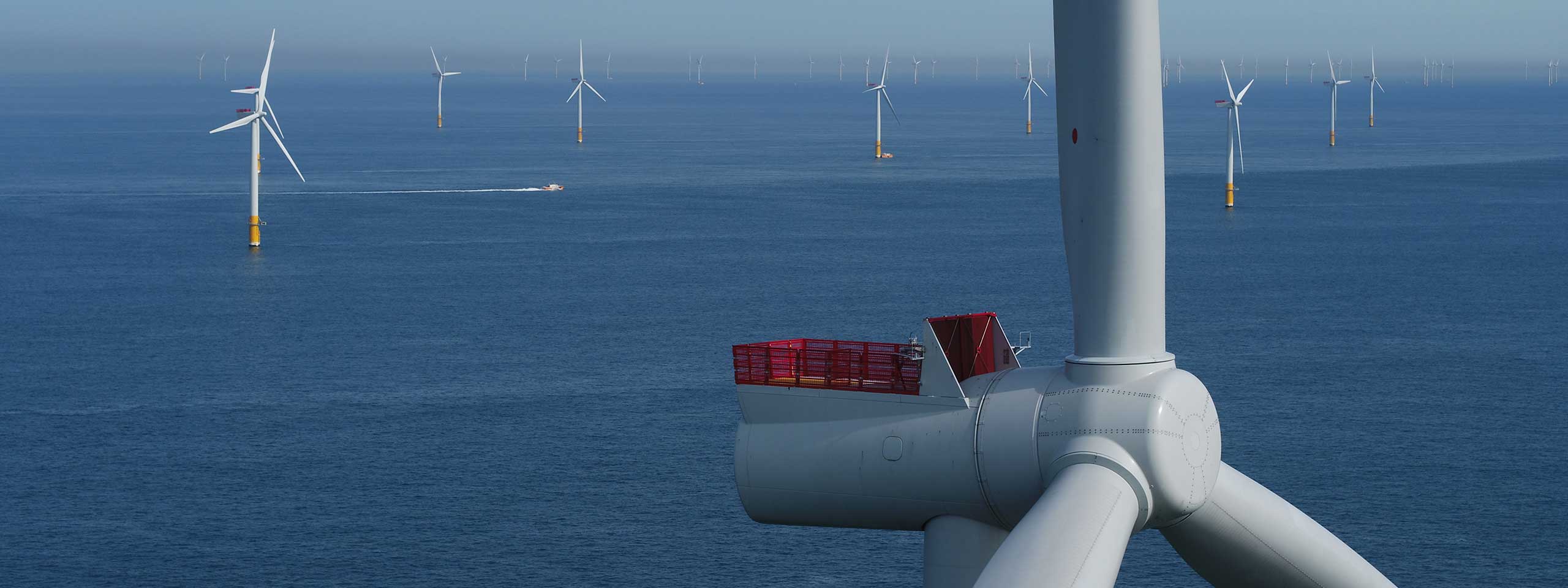 A wind turbine off the coast with several turbines in the background and a boat on the calm sea.