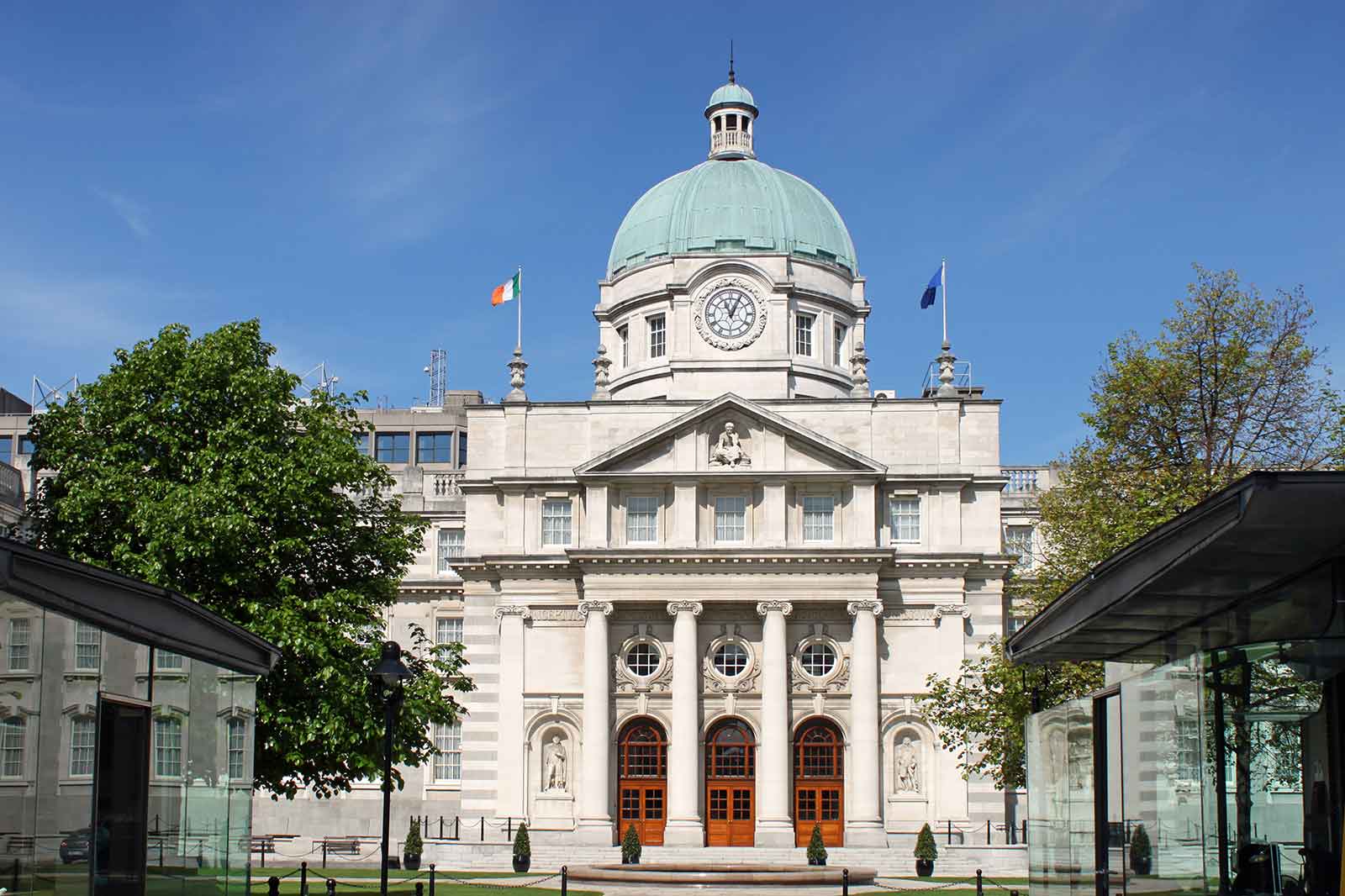 A grand building with a green dome, featuring pillars, flags, and a clear blue sky.