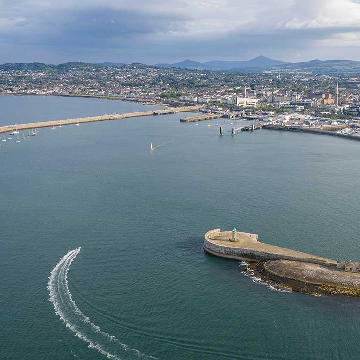 Aerial view of a harbour with a lighthouse, boats in the water, and a coastal town in the background.