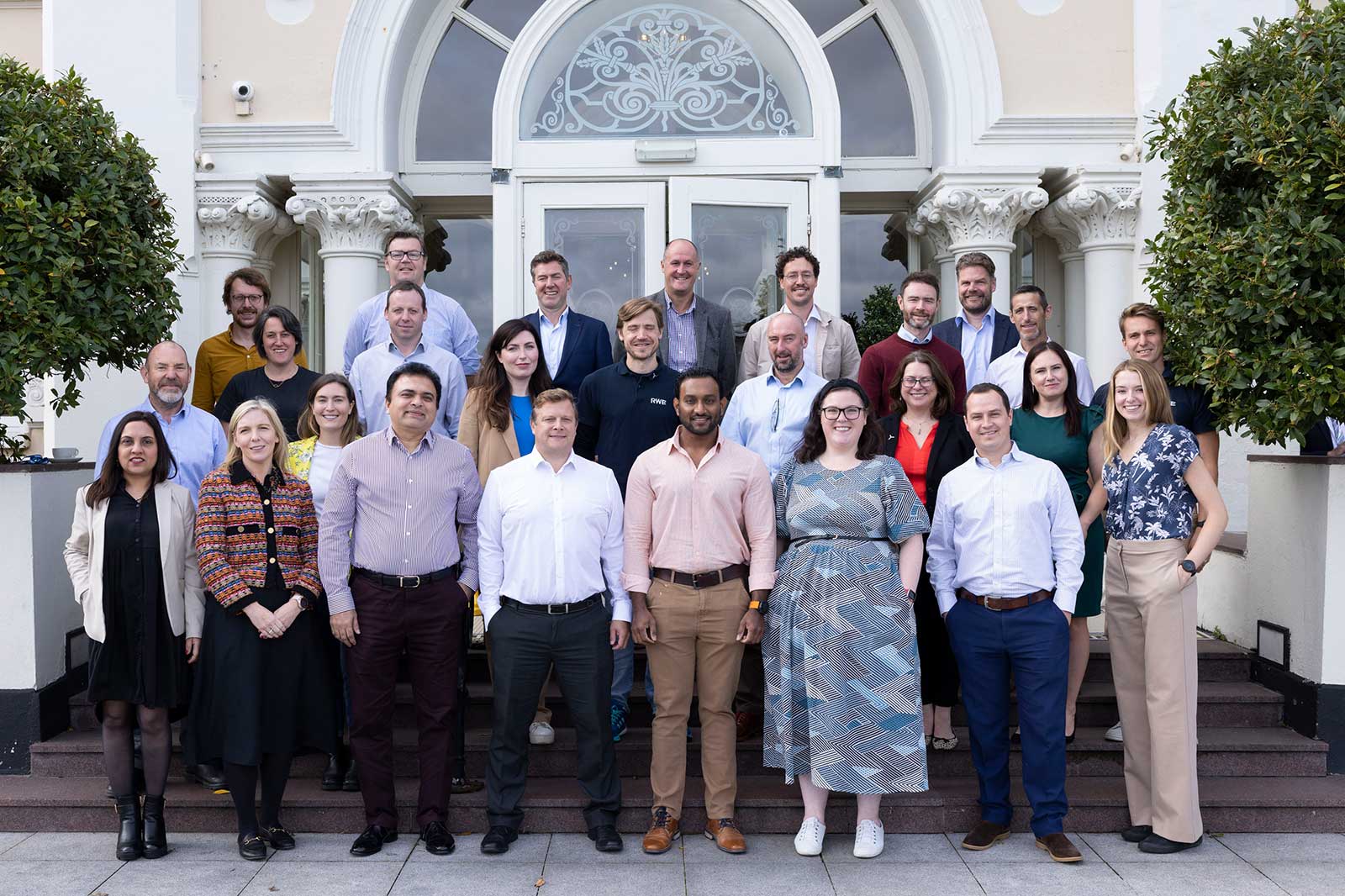 A group of individuals standing on the steps of a building, posing together outdoors, with diverse clothing styles and a decorative entrance.
