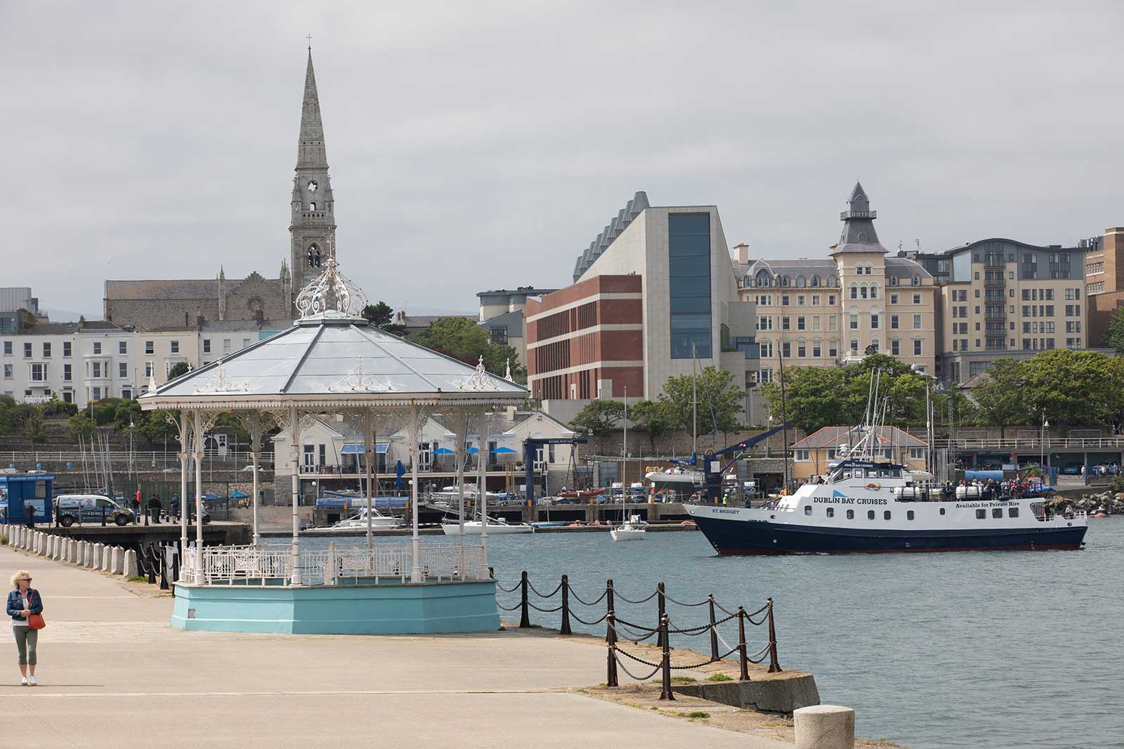 A seaside view featuring a bandstand, church spire, and a boat in the harbour, with buildings lining the shore.