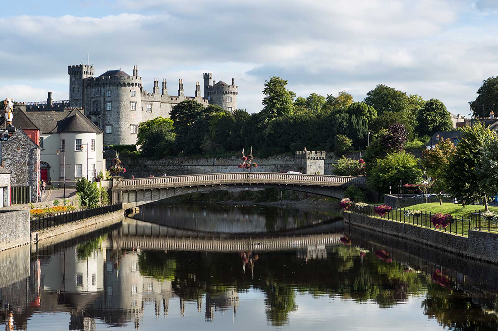 A scenic view of a castle beside a river, with a bridge and vibrant gardens reflecting in the water.