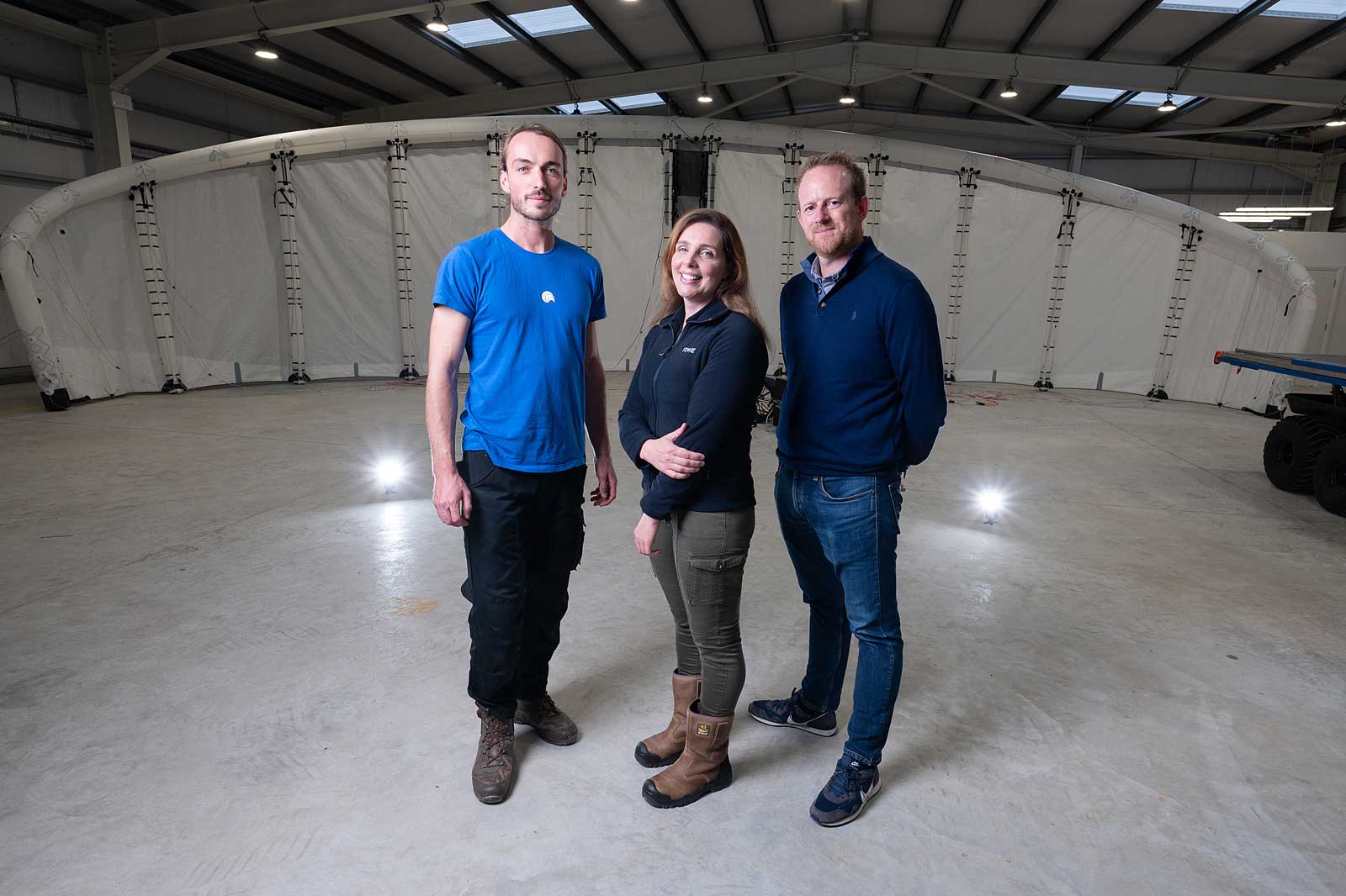 Three individuals stand in a spacious indoor area, with a large white structure behind them and subtle lighting.