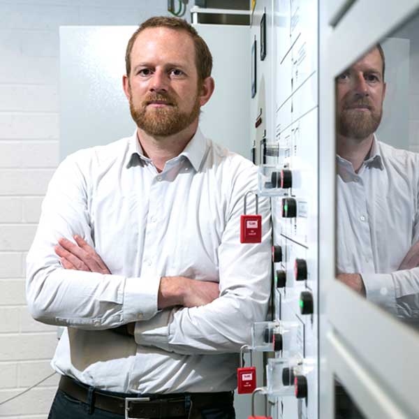 A man in a white shirt stands with arms crossed beside a control panel featuring various buttons and indicators, portrait of Cathal Hennessy,.