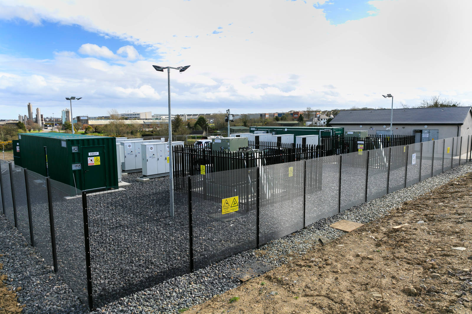 A fenced energy storage site with green containers and equipment, set against a cloudy sky.