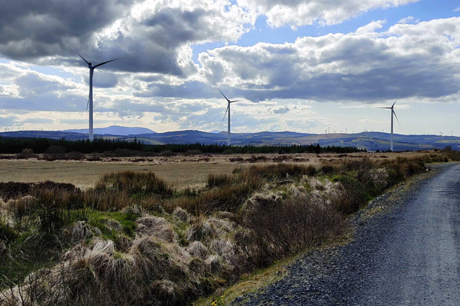 A scenic landscape featuring wind turbines in the distance, set against a cloudy sky and rolling hills.