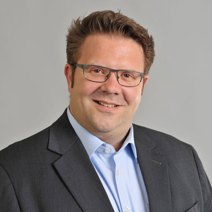 A person in a dark suit with a light blue shirt against a grey background. Portrait of Jan Peter Cirkel.